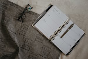 A flatlay image of a planner, eyeglasses, and pen on a textured fabric surface.