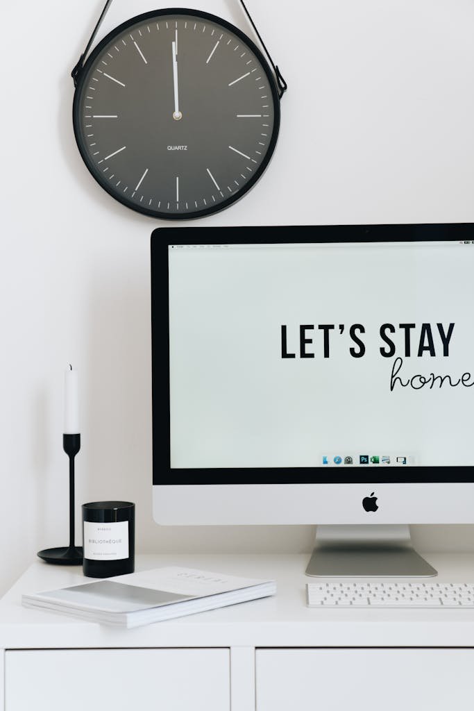 Clean and minimalist home office desk featuring a clock, monitor with 'Let's Stay Home' text, and decor.